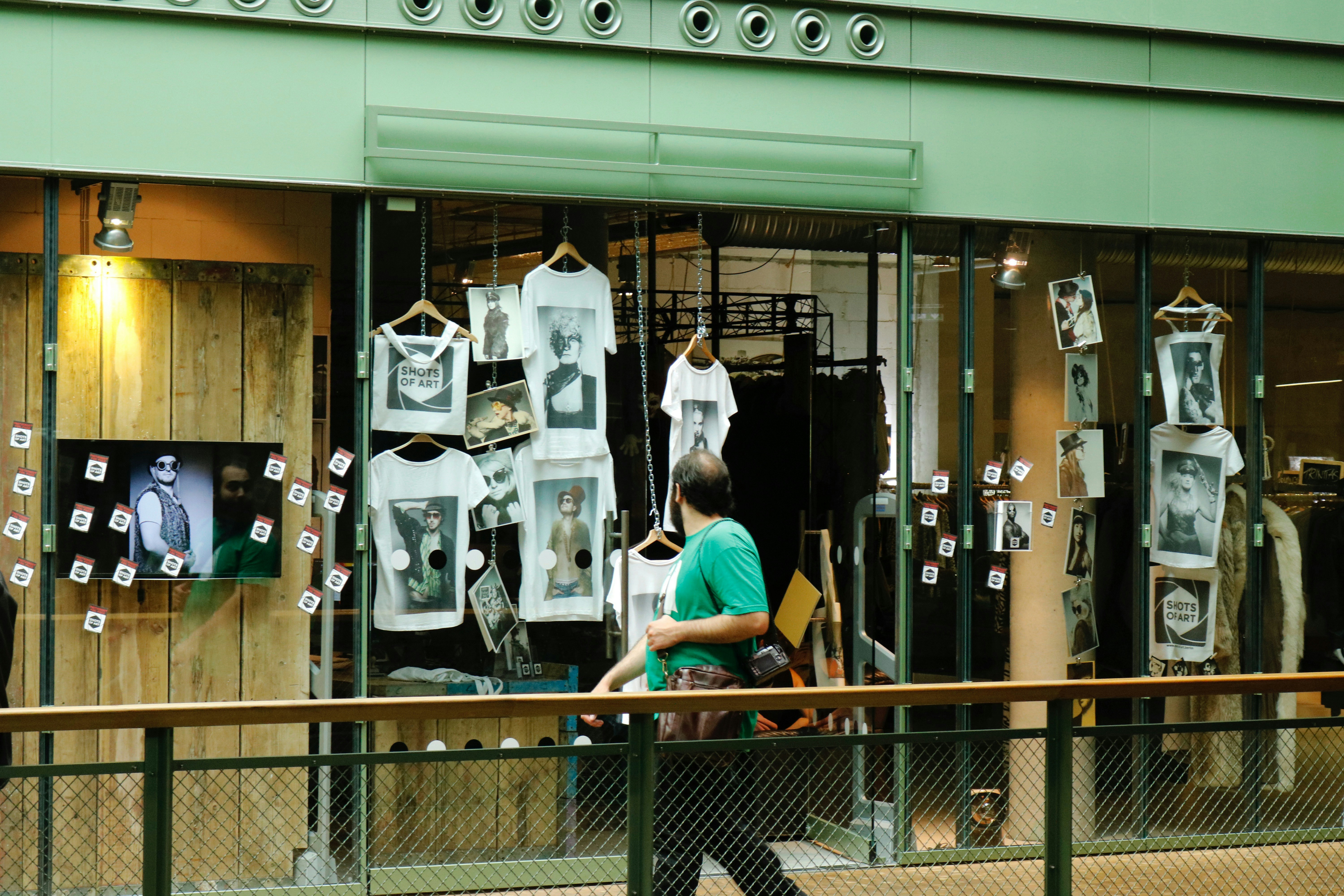 A shop window display featuring multiple white t-shirts printed with artistic black and white photographs. The storefront includes hanging t-shirts and several photographs attached to the glass. A television screen displays an image of a person in sunglasses. A person wearing a green shirt is walking past the display, holding a camera.