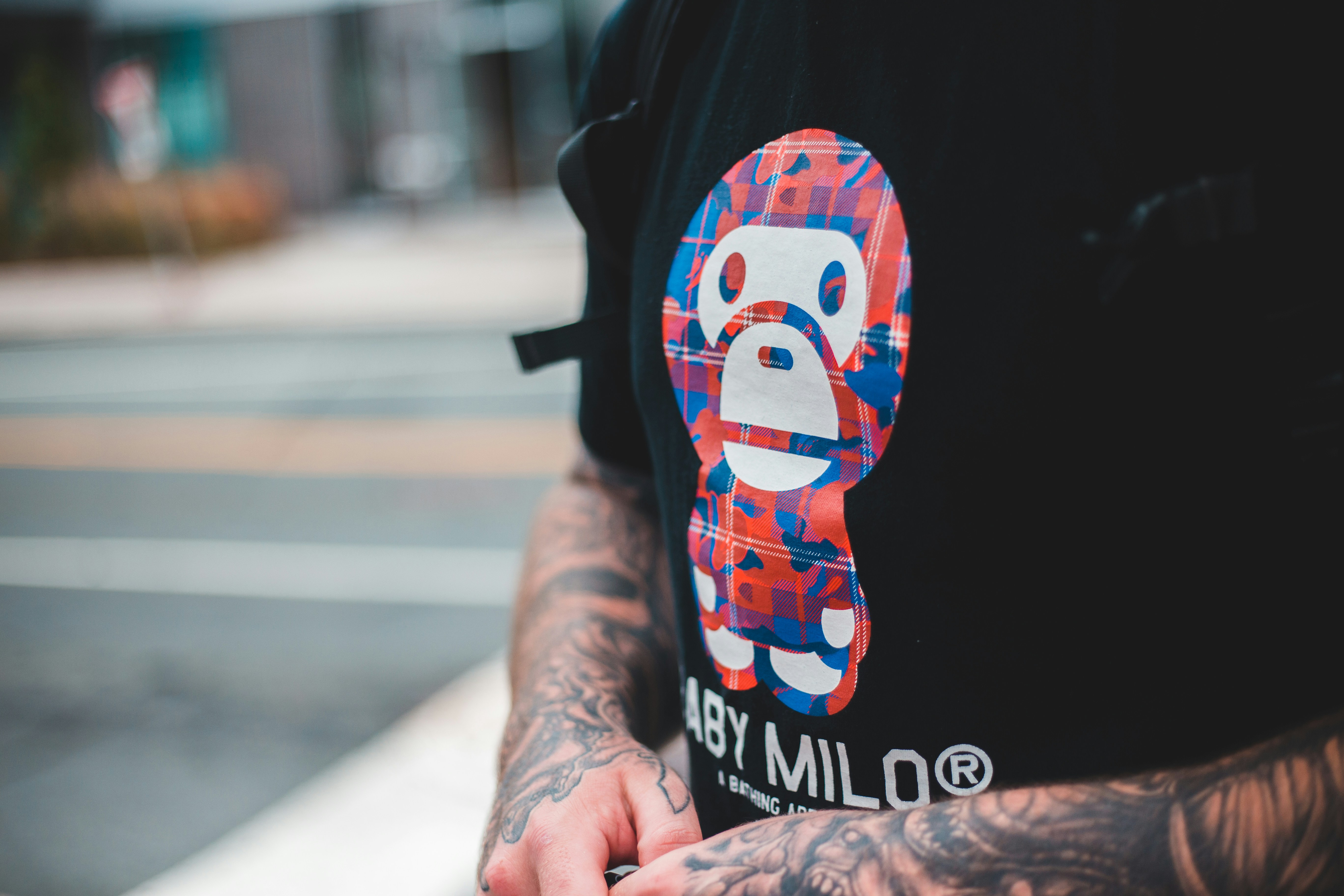 A person with tattooed arms is wearing a black T-shirt featuring a colorful graphic of a stylized character. The graphic consists of red, blue, and white colors arranged in a checkered pattern. The visible background is blurred, indicating an urban environment.