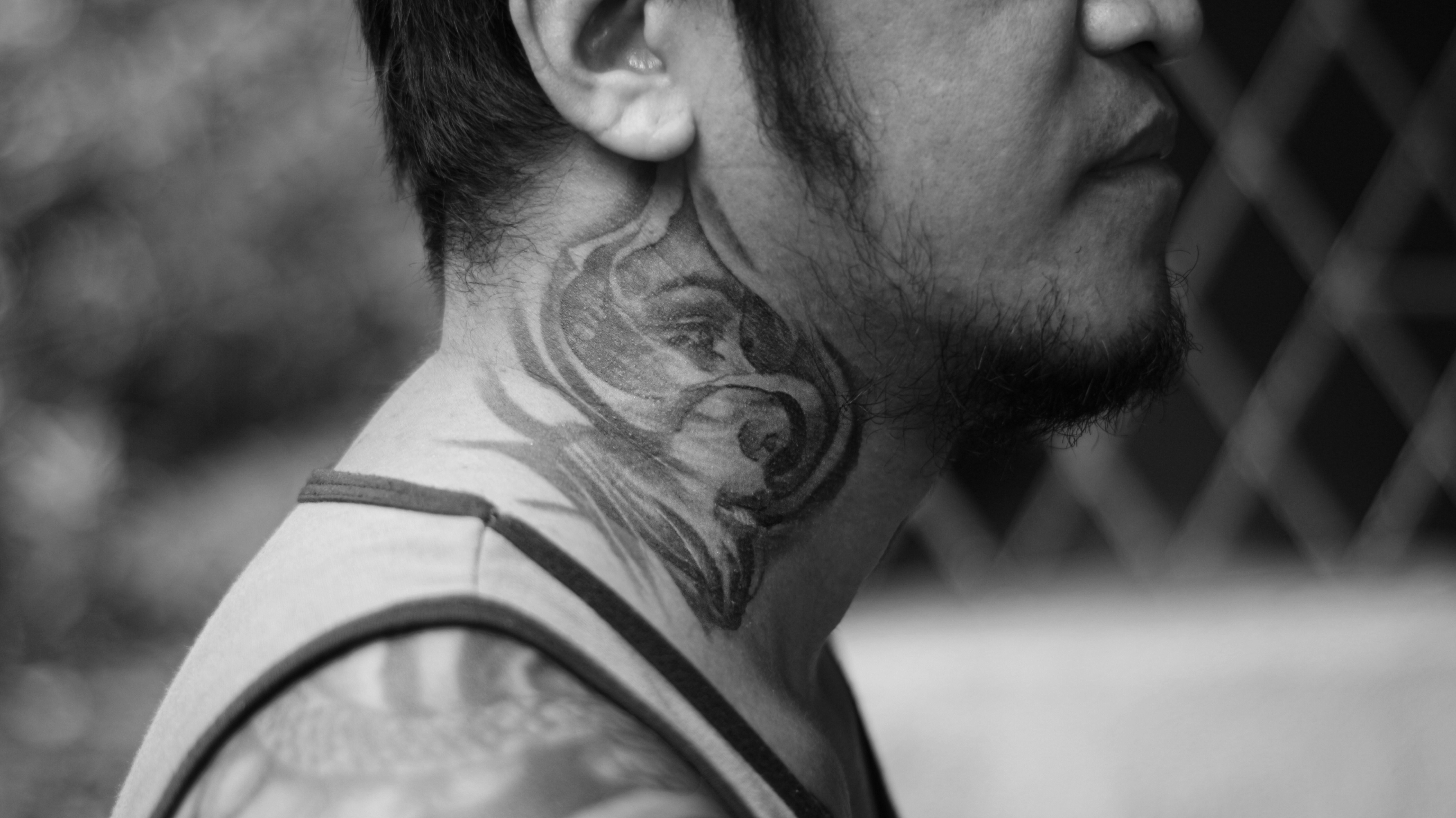 A close-up black and white photograph of a person with a detailed tattoo on their neck, extending to the shoulder. The tattoo design appears intricate and artistic. The person is wearing a sleeveless shirt, allowing a partial view of another tattoo on the arm.