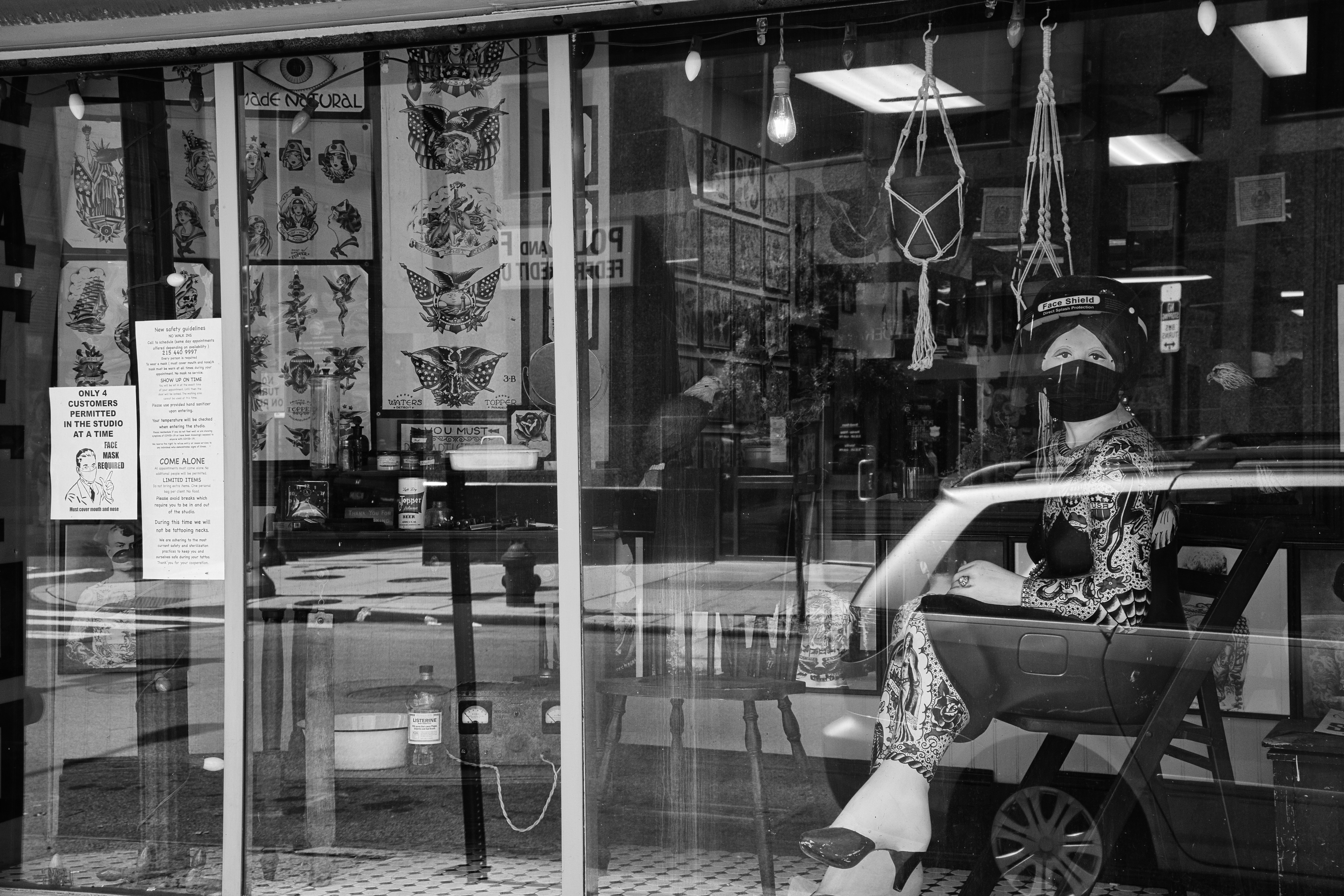 A black and white image showing a tattoo studio with large windows displaying tattoo designs. There is a mannequin or a person wearing a face mask and a face shield, sitting inside the shop. The interior includes hanging decorations and various objects related to tattooing. Signs on the window detail safety guidelines and restrictions.