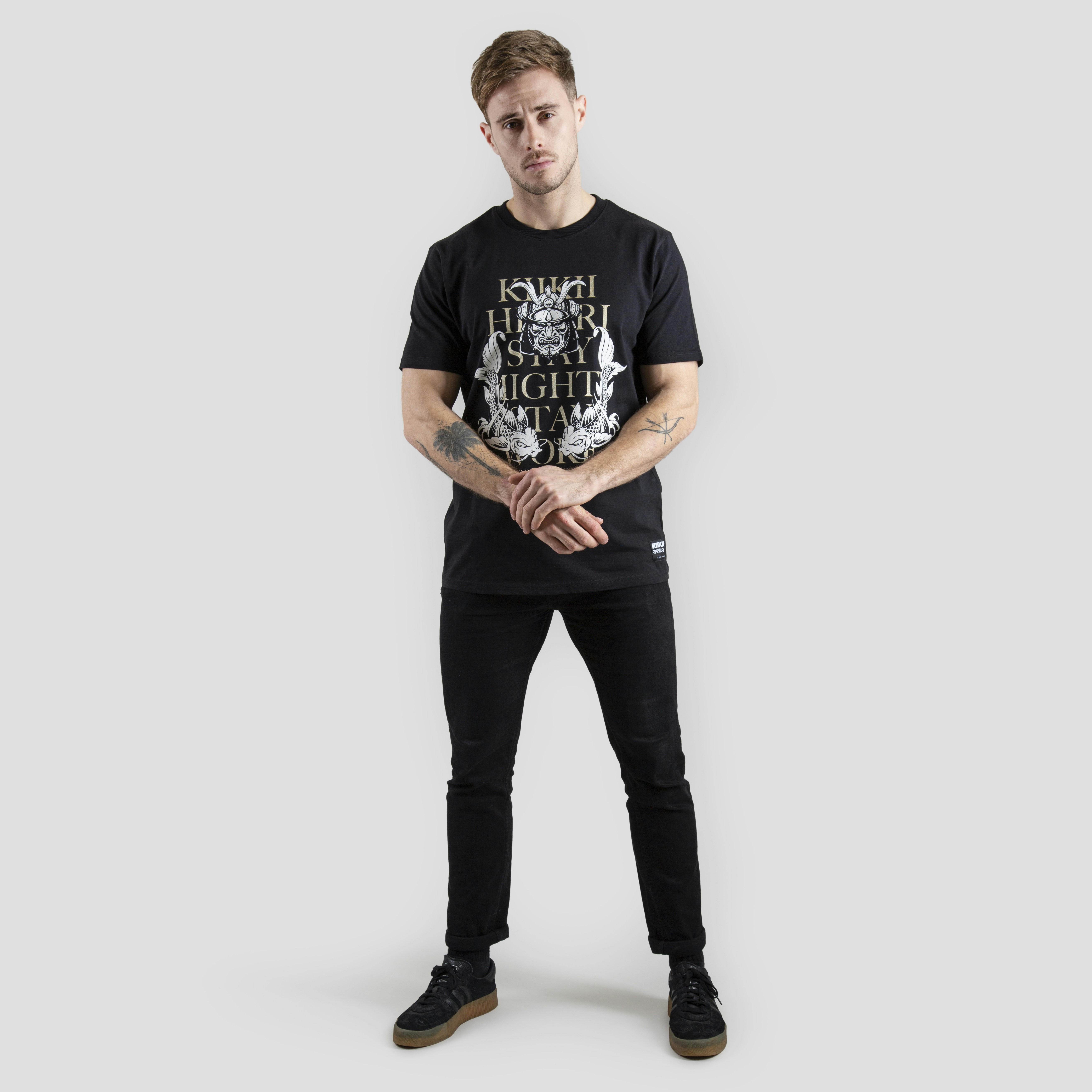 A person stands against a plain gray background wearing a black t-shirt with white and gold graphics. The design includes text and intricate patterns. The individual is dressed in black pants and black shoes with brown soles. Tattoos are visible on their forearms, and they have a neutral expression with arms loosely crossed.