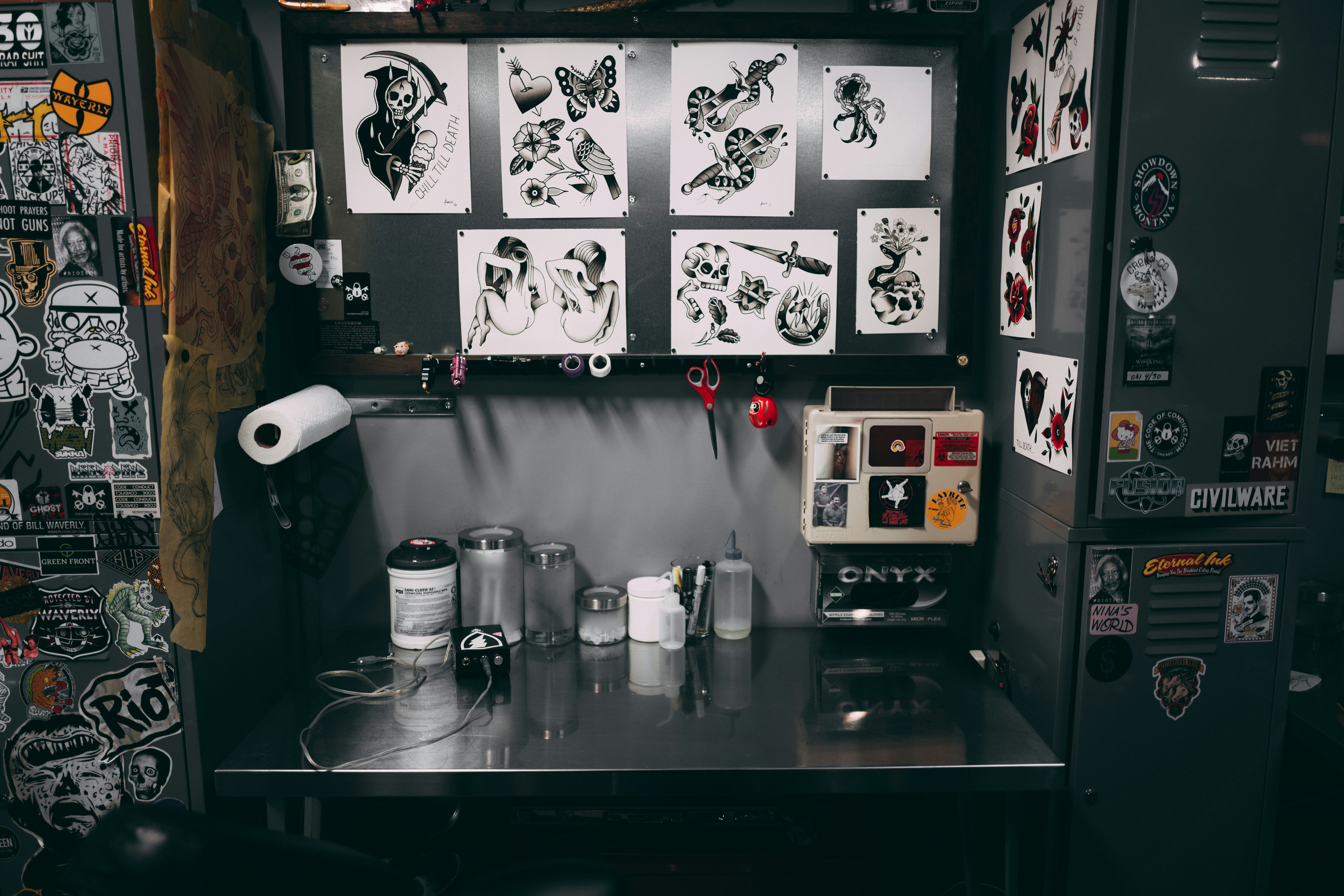 A tattoo workstation is cluttered with various items. The back wall displays numerous tattoo designs featuring hearts, daggers, skulls, and butterflies. The workbench holds ink bottles, paper towels, and various tattooing equipment. Stickers and artwork cover the surrounding surfaces, adding to the eclectic and artistic vibe.