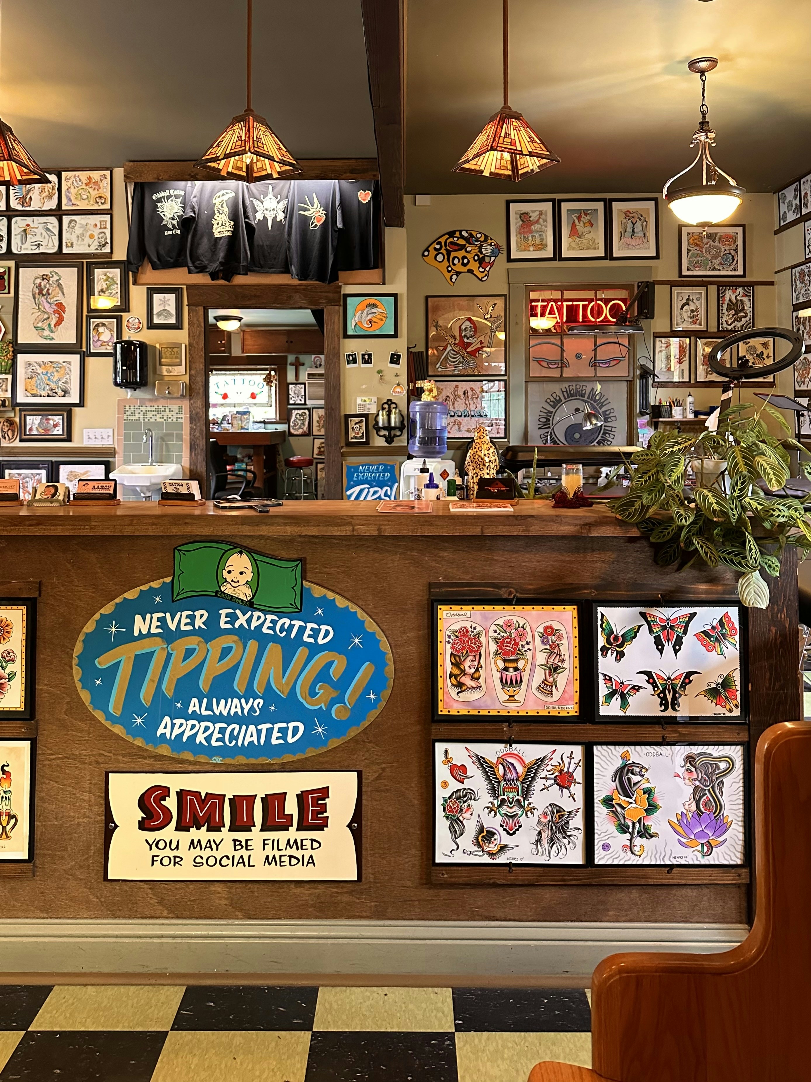 The image depicts the interior of a tattoo parlor. Artwork of various tattoo designs is displayed on the walls and a wooden counter. The counter features colorful signs related to tipping and social media. There are pendant lights with stained glass shades hanging from the ceiling, and a variety of framed pictures and designs provide a vibrant backdrop. A water cooler is present on the counter, and a collection of plants adds a touch of greenery.