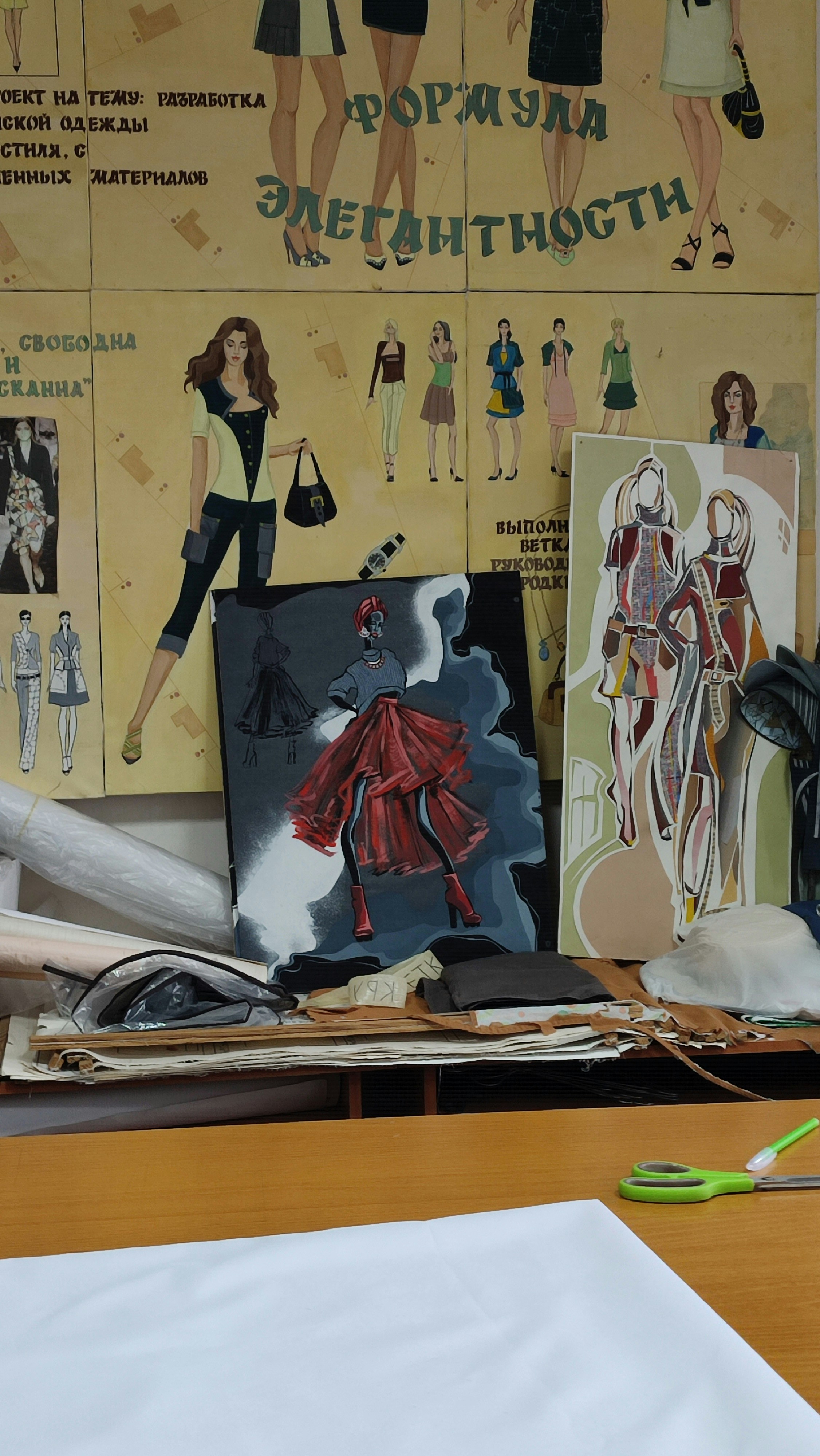 The image portrays a fashion design workspace with illustrated fashion sketches on a wall featuring various outfits. There are two canvases with vibrant artwork, one depicting a figure in a red dress. The surface is cluttered with papers, fabric rolls, and cutting tools.